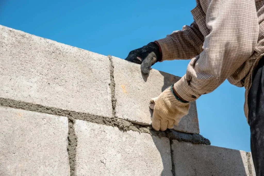 Construction worker building a concrete block wall, applying mortar between the blocks under a clear blue sky