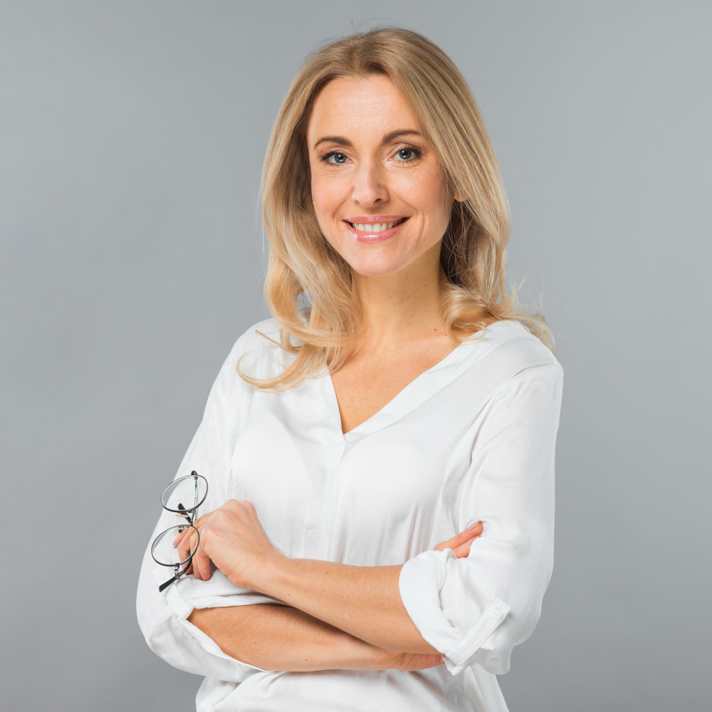 Smiling blonde woman with crossed arms holding glasses, wearing a white shirt on a gray background